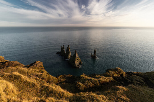 View of jagged dark sea stacks rise from the deep blue ocean under a streaked sky, from a golden grassy cliffside, Reykjav&Atilde;&shy;k, Reykjav&Atilde;&shy;kurborg, Iceland.