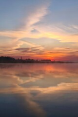 Obraz premium Serene sunset lake wrapped in soft mist rising from the water, glowing with rich orange, pink, and violet reflections, surrounded by gentle reeds and dark tree silhouettes under an expressive twiligh