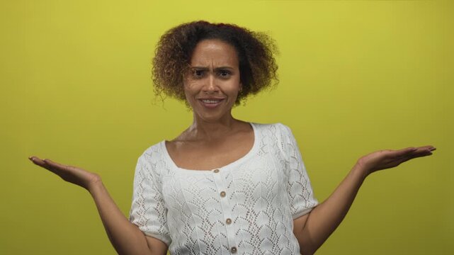 Woman waving hand with raised palm and puzzled facial expression in yellow studio wearing white knit top; confusion doubt questioning.