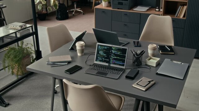 No people view of workplace of IT team with three beige chairs and two grey desks pushed together and set up with laptops, digital tablet, phones, notebooks and coffee cups