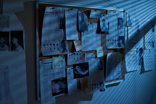 Corkboard covered with criminal profiles, mugshots, fingerprints, handwritten notes and investigation documents arranged for crime investigation in dimly lit room with shadow patterns