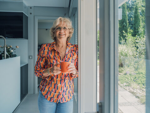 Smiling woman with orange cup standing in modern kitchen near window
