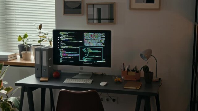 No people view of computer with source code on screen, wireless keyboard and mouse, notebook, lamp, stationery holder and two file folders on grey wooden desk in modern office