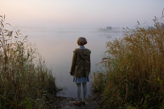 Woman with blonde short hair in a beige jacket by a foggy lake at sunrise