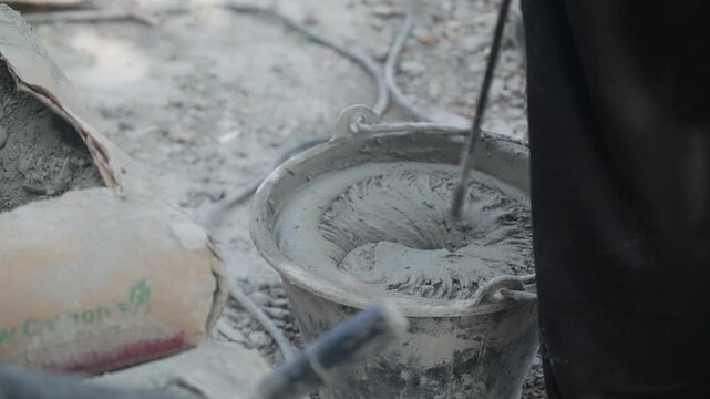 Construction worker mixing cement mortar in metal bucket at building site. Masonry preparation, renovation process and industrial material concept.