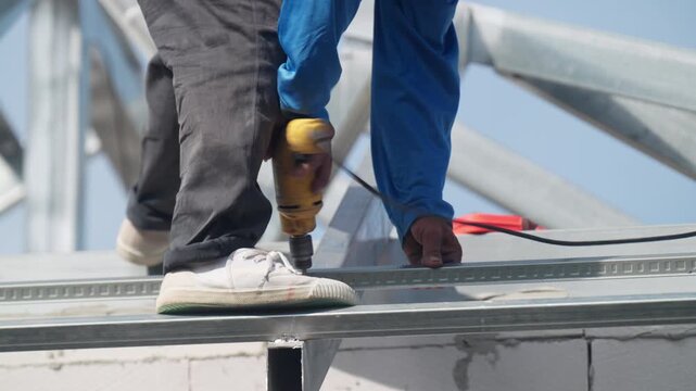 Construction worker using electric drill to fasten steel roof frame at building site. Industrial installation, structural engineering and roofing project concept.