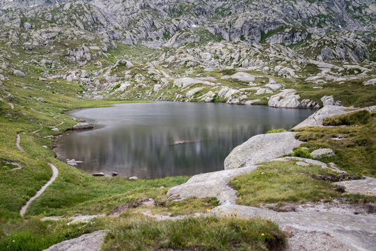 Peaceful alpine lake nestled among rocky mountain terrain and grassy slopes, with calm reflective water creating a serene wilderness atmosphere.