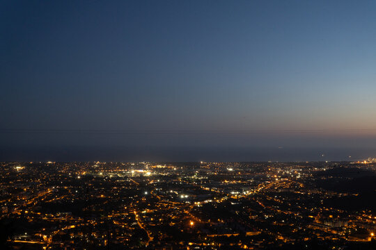 Panoramic cityscape at dusk with glowing streetlights and illuminated buildings stretching toward the horizon beneath a fading blue evening sky.