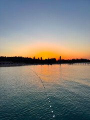 Fototapeta premium Early sunrise at Issyk Kul Lake in Kyrgyzstan. Warm golden sky reflecting in calm water with silhouettes of trees and pier on horizon.