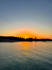 Naklejka premium Early sunrise at Issyk Kul Lake in Kyrgyzstan. Warm golden sky reflecting in calm water with silhouettes of trees and pier on horizon.