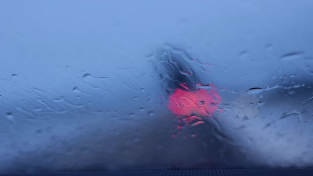Car ahead in rain seen through wet windshield. Red stoplight bokeh scene.