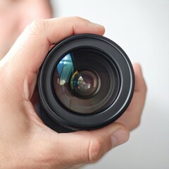 Close up of a hand holding a black camera lens with reflections in the glass against a white background
