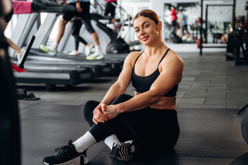 Fototapeta premium Female fitness enthusiast in black athletic wear sits on gym floor, smiling, with treadmills and other gym-goers visible in the background
