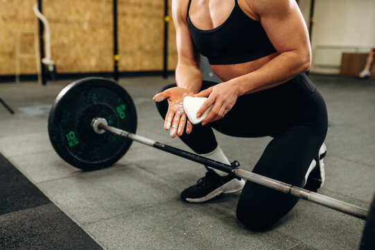 Female athlete preparing for weightlifting session, applying chalk to hands while kneeling beside a barbell on gym floor with weights visible in the background
