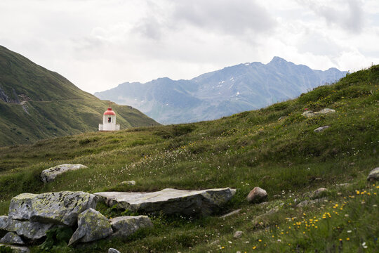 Scenic alpine landscape with rolling green hills, rugged mountain peaks. A small white structure with a red dome stands alone in the meadow, adding a focal point to the peaceful highland scenery.