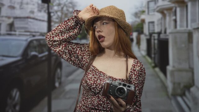 Redhead woman holding vintage camera, hand to hat and exposed shoulder, standing on a narrow street with parked car and stone building; melancholy wanderlust nostalgia.