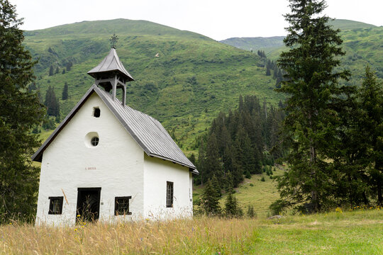 Charming white mountain chapel surrounded by lush green hills and tall pine trees. simple architecture contrasts the natural alpine scenery, creating a serene and pastoral countryside setting.