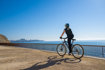 Obraz premium Rear view of female cyclist pedaling along Mediterranean waterfront trail with expansive sky and ocean backdrop, symbolizing exploration, independence and active outdoor lifestyle.
