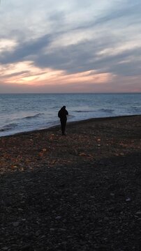 Lonely man walking along a deserted pebble beach during a colorful sunset. Calm sea waves gently wash ashore as he enjoys a quiet, introspective moment in nature