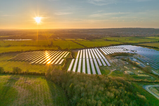 Aerial view of solar panels reflecting the golden sunset light amidst the verdant fields of Cowley Baldon Green Solar Farm, Cowley, Oxfordshire, United Kingdom.