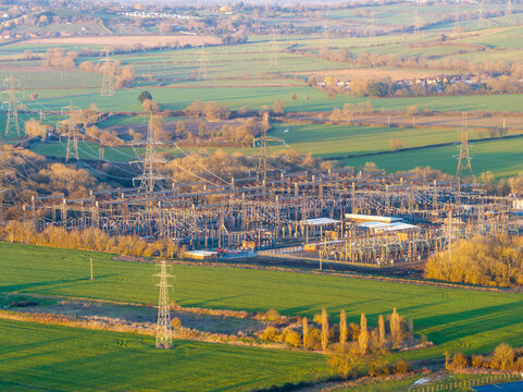 Aerial view of a sprawling electrical substation amidst vibrant green fields, pylons marching across the landscape under a soft, golden light, Cowley, United Kingdom.