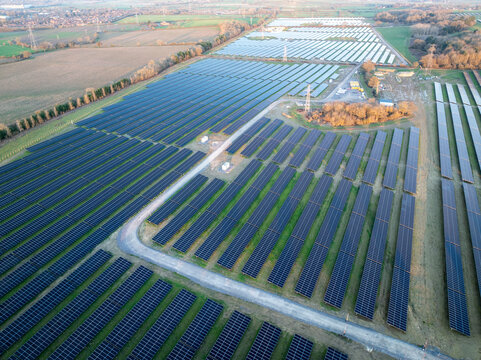 Aerial view of a vast solar farm, with symmetrical rows of dark panels contrasting with the surrounding golden fields and light green trees, Cowley, Oxfordshire, United Kingdom.