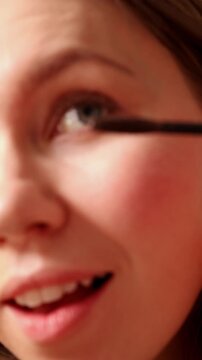 A vertical macro shot with a focus on a gold mascara tube and brush. A young woman's face is blurred in the background as she prepares to apply makeup