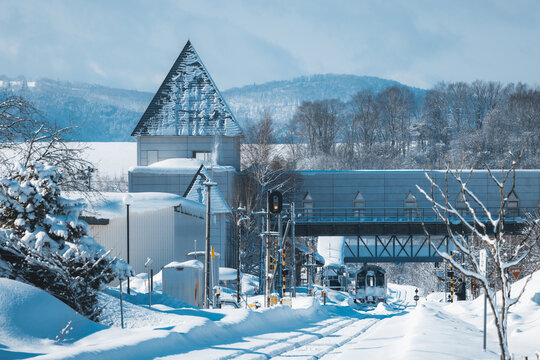 View of a snow-laden railway track leading towards a distant train under a covered bridge, with snow-covered trees and hills in the background, Biei, Hokkaido, Japan.