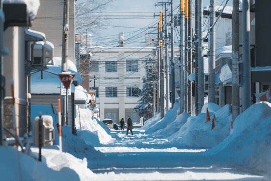 View of a snow-laden street, flanked by towering snowbanks and utility poles under a clear sky, where two figures walk in the distance, Biei, Hokkaido, Japan.