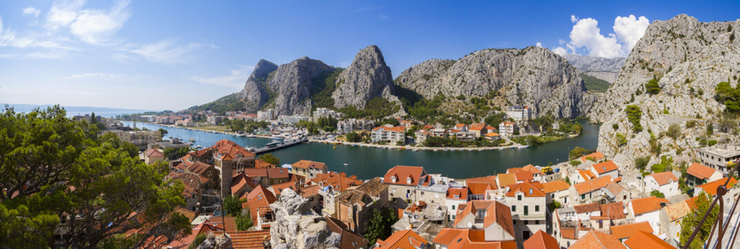 View of terracotta rooftops cascading down to the shimmering river framed by colossal mountains under a bright blue sky, Omis, Split-Dalmatia County, Croatia.