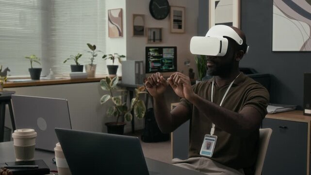 Black young male quality assurance engineer evaluating software product using VR headset while sitting at office desk