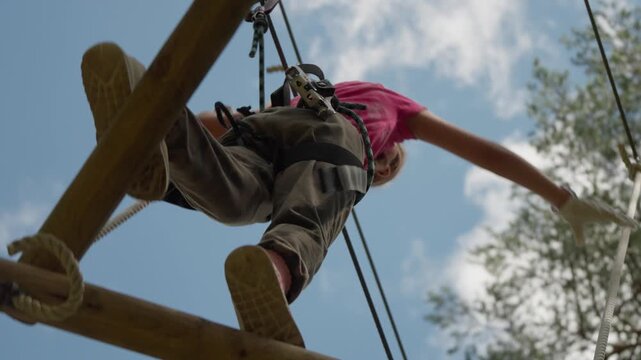 Person navigating high ropes course at adventure park in Stockholm. Individual wearing safety harness balances on wooden beam during summer outdoor activity. Low angle view against blue sky.