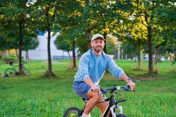 Naklejka premium Smile guy is riding a bicycle on a path in a grassy park on a sunny afternoon. He is wearing shorts and a sweatshirt and looks happy enjoying the outdoors
