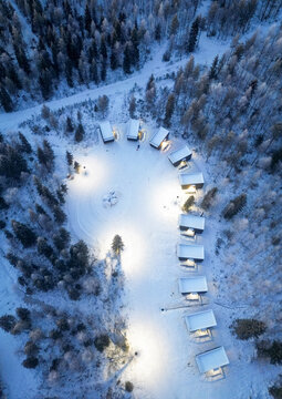 Aerial view of snow-covered cabins nestled amidst a forest, lights glowing warmly against the cold twilight, Rovaniemi, Finland.