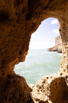 View of the azure sea through a craggy cave opening, where sunlight dances on the water's surface, contrasting with the cave's textured walls, Gruta da Boneca, Faro, Portugal.