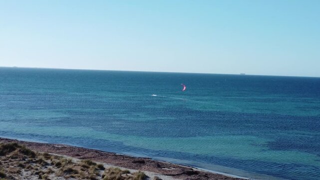 Stunning aerial footage showing a lone kitesurfer navigating the vast turquoise and deep blue ocean on a bright, sunny day, with a sandy coastline visible in the foreground