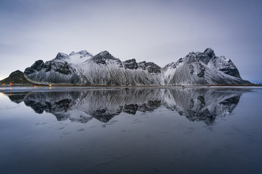 View of stark, snow-dusted mountains mirroring flawlessly on the dark, still water under the predawn sky, creating a serene yet dramatic landscape, Hofn, Sveitarfelagid Hornafjordur, Iceland.