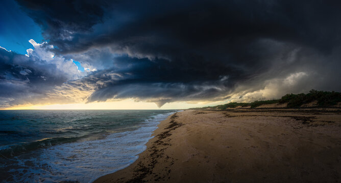 View of ominous dark storm clouds roll in over the sandy beach and turbulent sea, contrasting with the golden light, Taranto, Apulia, Italy.