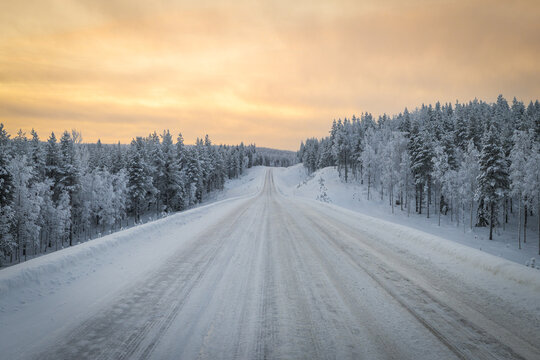 View of a long road stretching through a snow-covered forest under a soft, warm sky, creating a serene winter landscape, Ruka, Pohjois-Pohjanmaa, Finland.