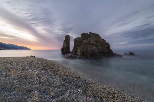 View of a rugged coastline where the sea gently laps against a shingle beach under a sky painted with hues of dawn, Monterosso al Mare, Liguria, Italy.