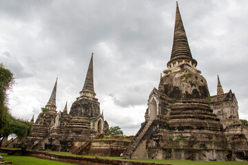 Obraz premium The three iconic restored chedis (stupas) at Wat Phra Si Sanphet in Ayutthaya, Thailand. The massive, bell-shaped structures stand in a row on a raised platform covered in green grass. Cloudy day.