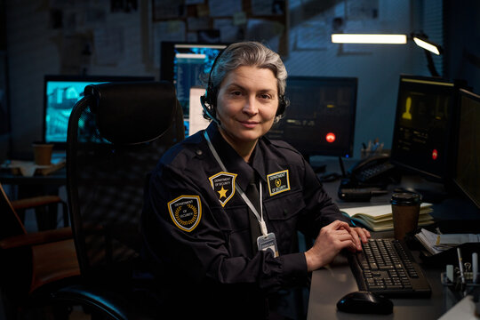 Portrait of middle aged Caucasian woman police officer sitting at desk, wearing headset, working at computer in control room with multiple monitors and documents visible in background