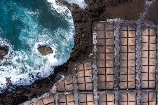 Aerial view of the contrasting turquoise waves crash against the dark, jagged coastline next to the geometric salt pans, Santa Cruz de La Palma, Spain.