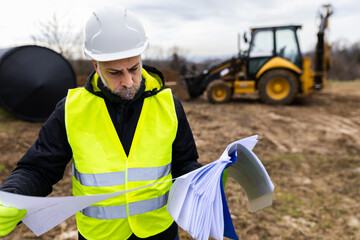Engineer analyzing blueprints with excavator in background