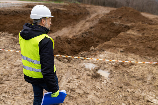 Worker inspects deep excavation site while holding plans in hand during a cloudy day