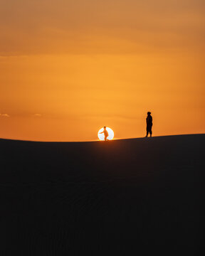 View of silhouetted figures stand amidst the ochre glow of the setting sun, casting long shadows across the rippled dunes, Da'ira, Yemen.