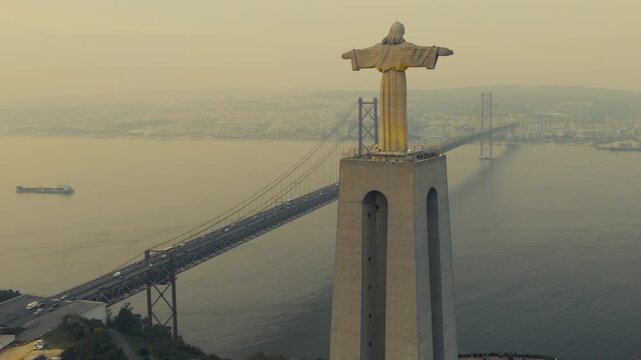 Drone view of the Christ the King monument in Almada, Portugal, with the 25 de Abril Bridge spanning the Tagus River and Lisbon visible in the background.
