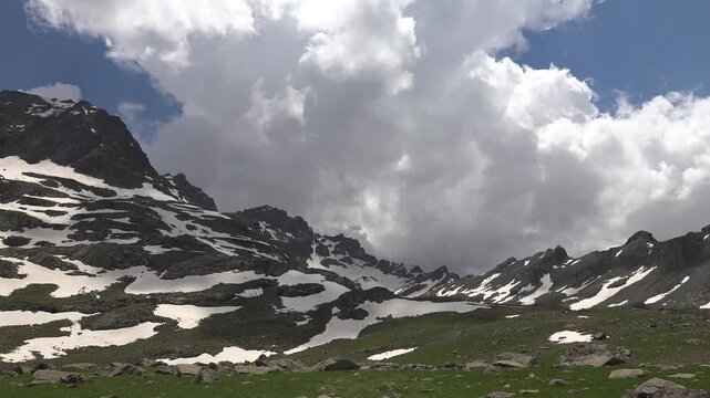 Timelapse clouds drift above snow patches on Rocky Mountains, spring alpine meadow, USA. Fast moving sky rolls past rugged ridges and green grassland as melting drifts fade in America.