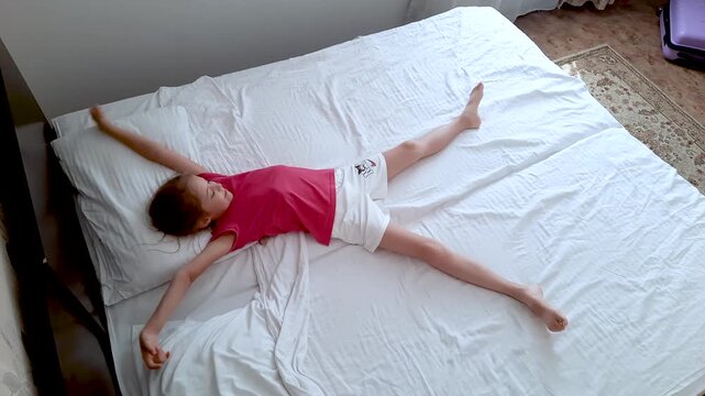 A happy girl lies on a bed, stretches, and waves her arms and legs, viewed from above.
