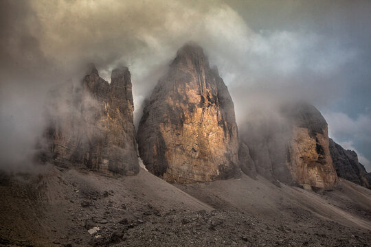 View of jagged peaks pierce through swirling clouds, bathed in ethereal light above the rocky terrain, a serene yet dramatic landscape, , Tre Cime di Lavaredo, Veneto, Italy.